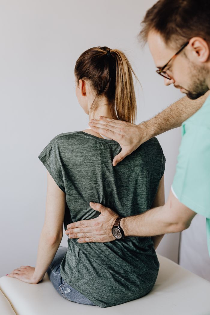 From above of crop chiropractor in eyeglasses and uniform checking up back of slim faceless female patient in casual wear sitting on examination couch in clinic